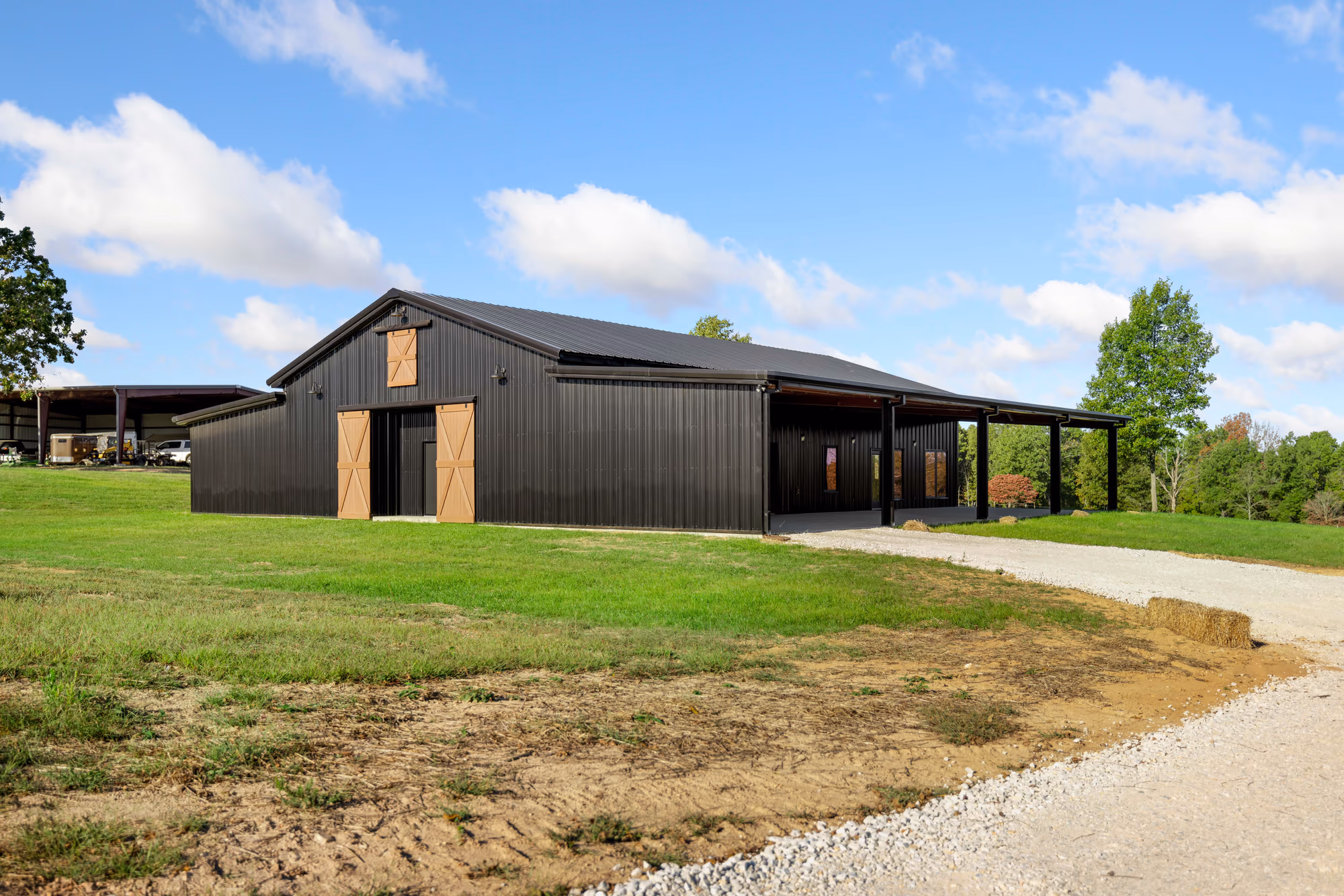 A black metal barndominium with a covered lean-to porch running its length, standing on a mown rural lot at the end of a gravel drive