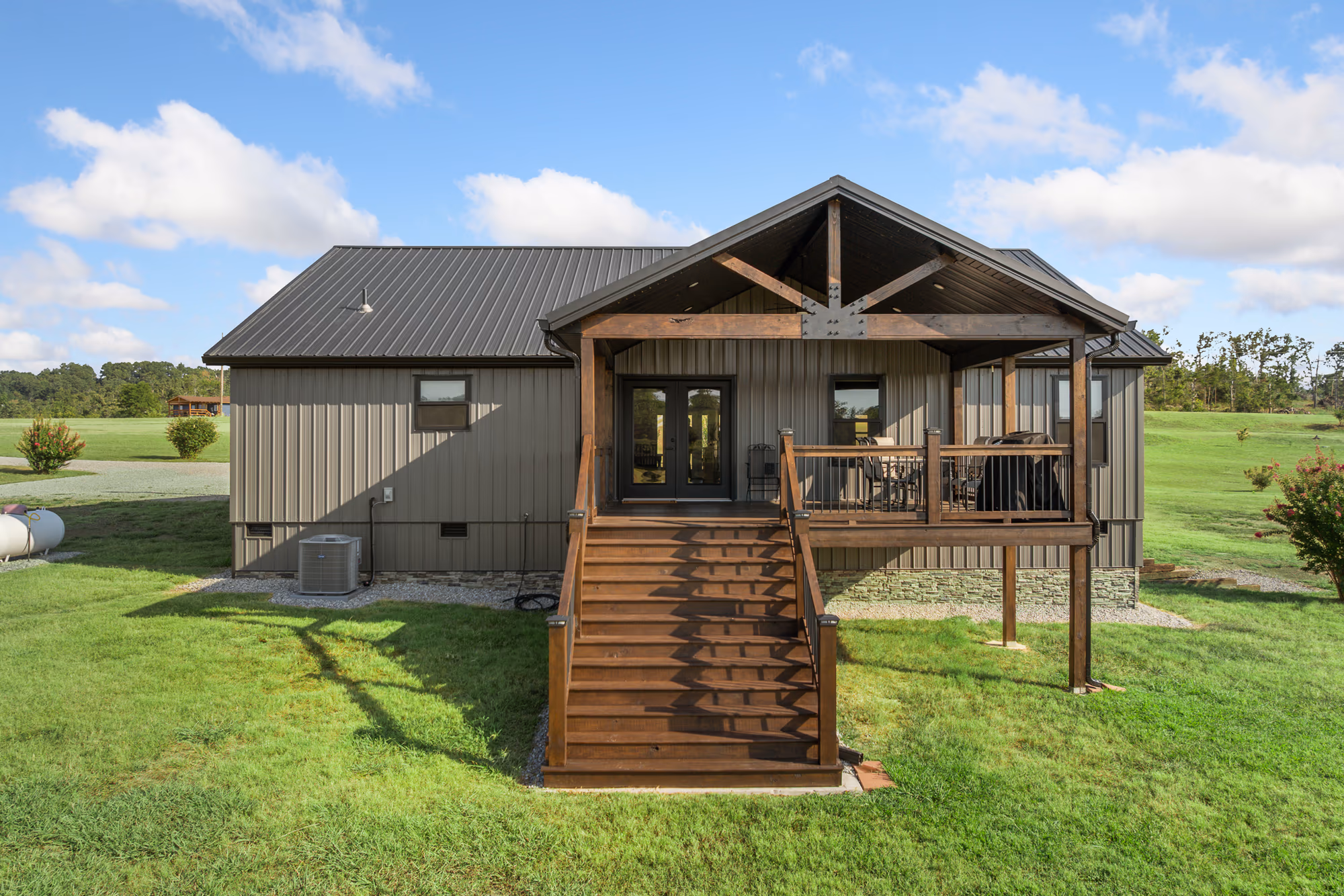 A barndominium in olive-tan vertical metal siding under a dark standing-seam roof, its entry sheltered by a stained timber gable truss above a wood stair and a stone wainscot