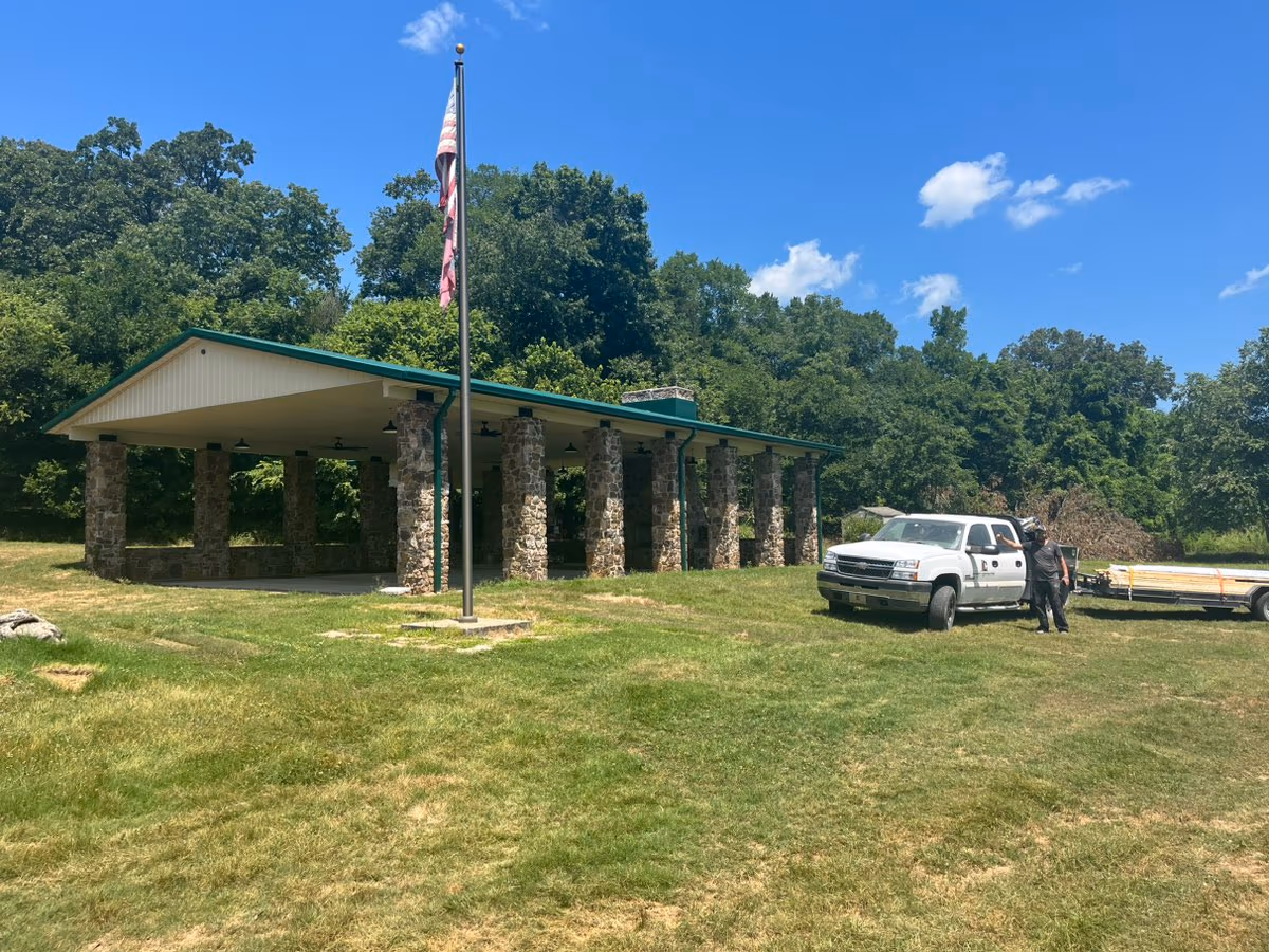 The Arkansas Game & Fish pavilion — a long open shelter carried on stacked-stone columns under a green standing-seam metal roof, with a flagpole on the lawn in front