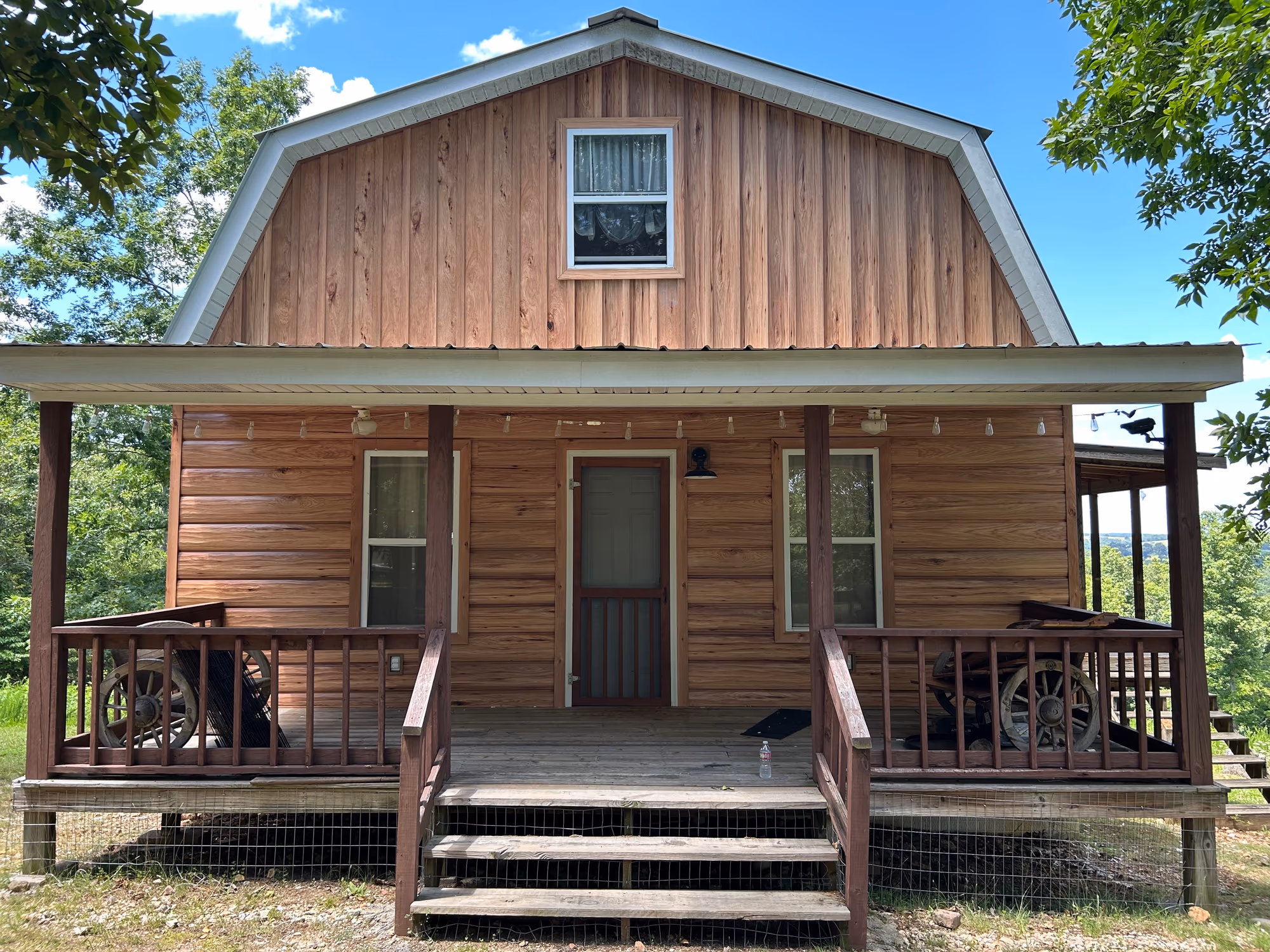 A gambrel-roofed cabin clad in TrueLog metal siding that reads as stacked timber, with a covered porch, wagon-wheel railings and a metal roof