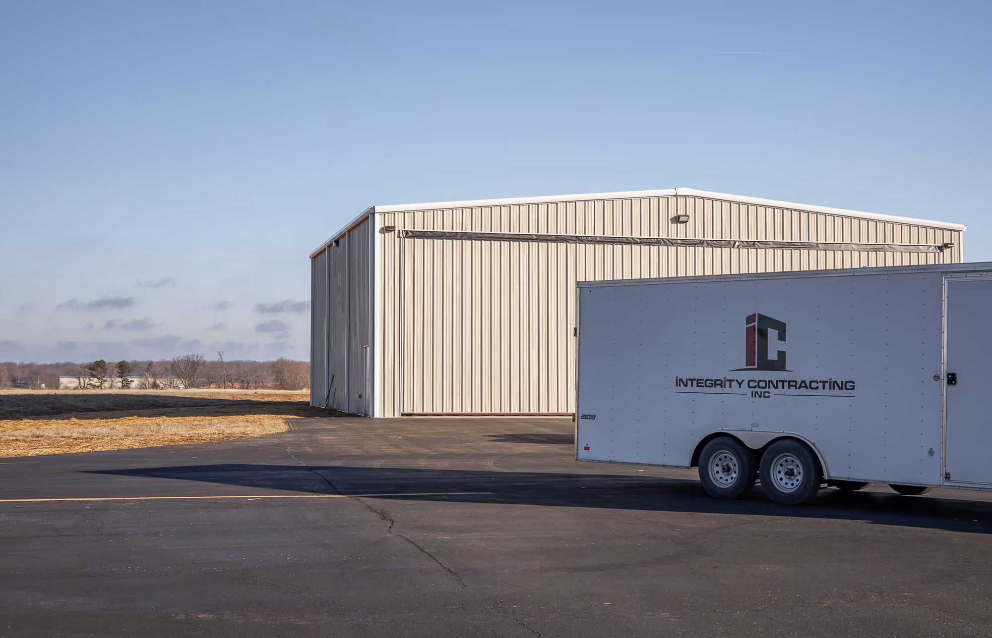 A cream steel aircraft hangar on a level apron, with a branded Integrity Contracting equipment trailer parked in front of it