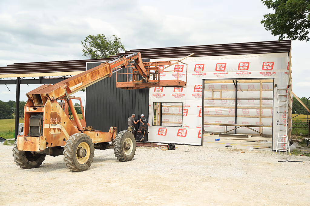 An Integrity Contracting crew hanging dark metal siding from a telehandler basket, over a house-wrapped timber frame with the red-iron eave already in place